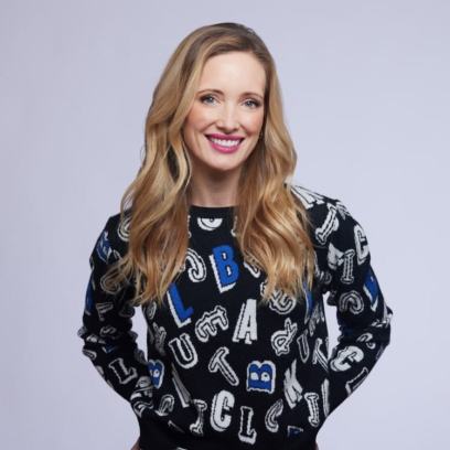 Jessica Reid Sliwerski stands in front of a grey backdrop wearin ga sweater covered in letters of the alphabet