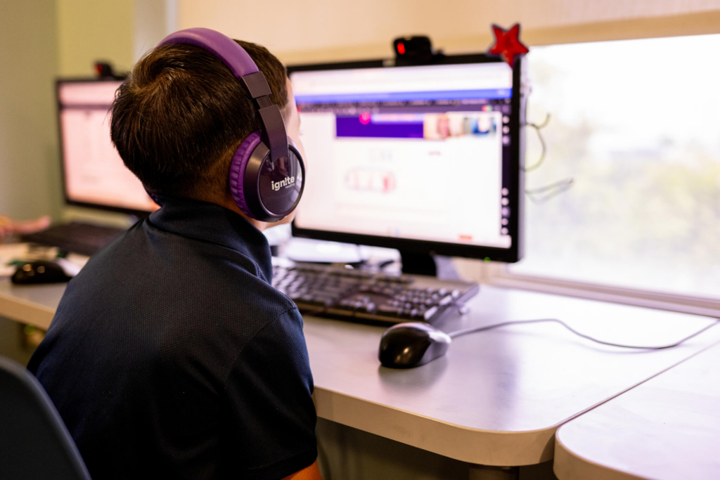 two 2nd grade boys sit in a classroom wearing headphones and working on computers with their online tutors