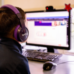 First Grade student wearing headphones talks to his virtual tutor while facing computer screen