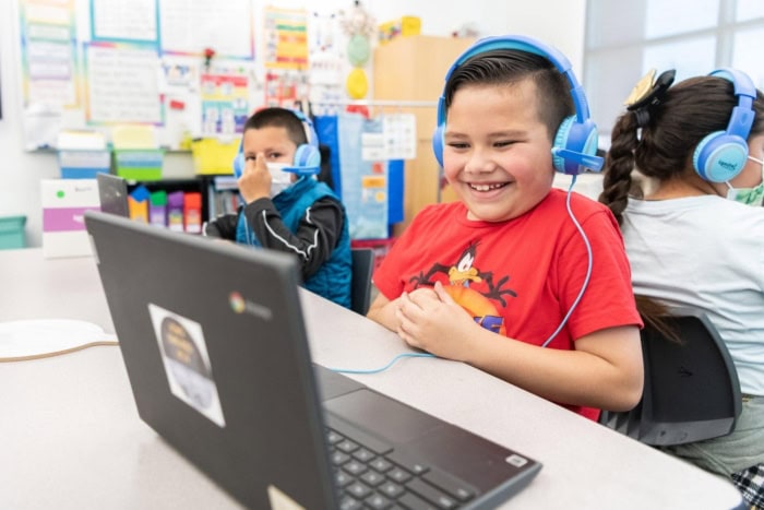 two 2nd grade boys sit in a classroom wearing headphones and working on computers with their online tutors