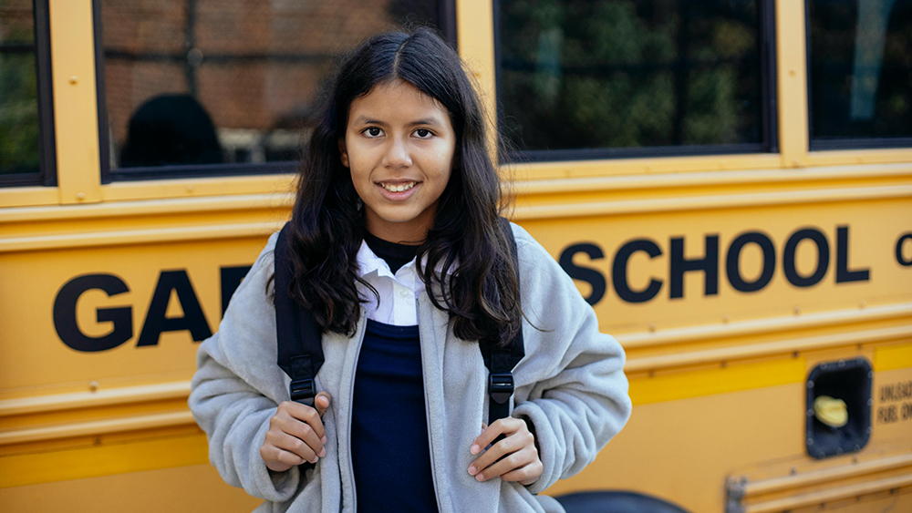 Multilingual Learner stands in front of a School Bus