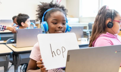 a 1st grade student holds up a whiteboard with the word hop written on it so her online tutor can see it through the computer screen