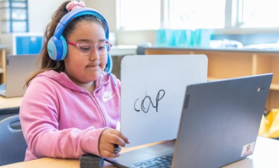 An elementary student holds a whiteboard with the word cap on it so her reading tutor can see it