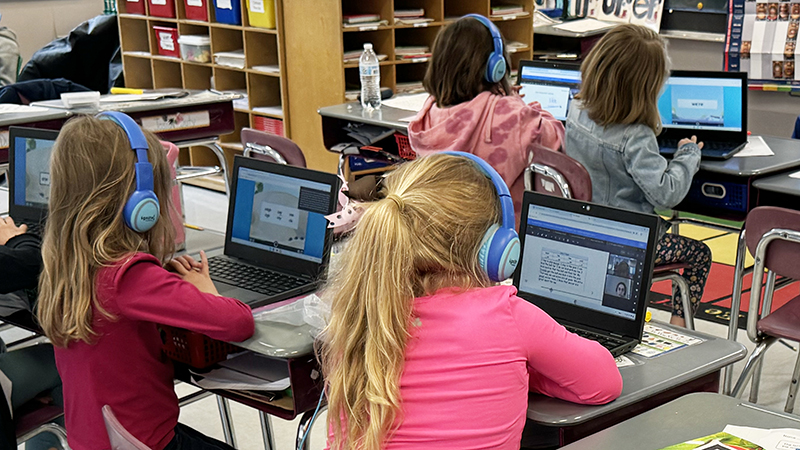 4 students in a classroom are seen in their high dosage tutoring sessions. The students are all wearing headphones and facing their online tutors on a computer screen.